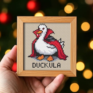 Op de afbeelding: Kruissteekkunstwerk in een houten lijst. Het ontwerp toont een witte eend met een rode cape en het woord "DUCKULA" in zwarte letters. De eend heeft zwarte en grijze accenten en oranje voeten. De achtergrond is een lichtbeige stof.