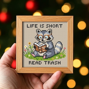 Könnte beinhalten: Ein gerahmtes Kreuzstichbild mit einem Waschbären, der ein Buch liest, mit dem Text "LIFE IS SHORT READ TRASH". Der Waschbär ist grau und weiß, trägt eine Brille und sitzt in einem Feld mit grünem Gras und Blumen. Der Rahmen ist aus hellbraunem Holz.