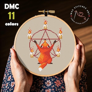 May include: Cross-stitch pattern featuring a stylized orange fox within a pentagram, surrounded by candles. The design is set against a beige fabric, held in a wooden embroidery hoop. Text includes "DMC 11 colors" and "MAX STITCHES ART CROSS STITCH PATTERNS".