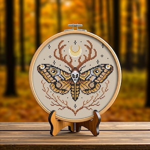 May include: Cross-stitch embroidery hoop with a beige fabric background featuring a detailed design of a moth with a skull head, antlers, and a crescent moon. The moth has brown, yellow, and black wings with white accents.