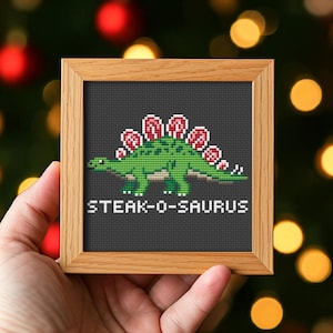 Puede incluir: Un bordado a punto de cruz enmarcado que representa un estegosaurio verde con detalles rojos. El texto "STEAK-O-SAURUS" está bordado debajo. El marco es de madera marrón claro.