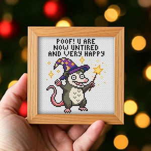 May include: A square wooden frame containing a cross-stitch embroidery of a gray and white opossum wearing a purple wizard hat with yellow stars. The opossum holds a yellow star wand, with the text POOF! U ARE NOW UNTIRED AND VERY HAPPY.