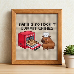 May include: Cross-stitch art in a wooden frame. The design features a red oven with a tray of cookies and a brown capybara wearing a chef's hat. Text reads: "BAKING SO I DON'T COMMIT CRIMES."