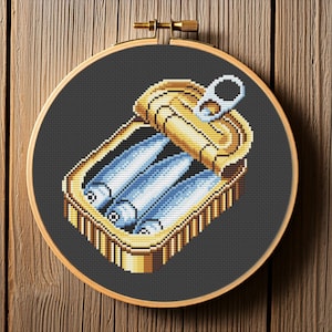 May include: A cross-stitch embroidery hoop with a design of a can of sardines. The can is open and the sardines are visible inside. The can is gold and the sardines are blue and white.