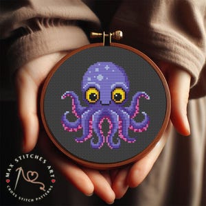 Könnte beinhalten: Ein lilafarbener Oktopus mit gelben Augen und rosa Tentakeln ist auf einem dunkelgrauen Stoff in einem runden Stickrahmen gestickt.