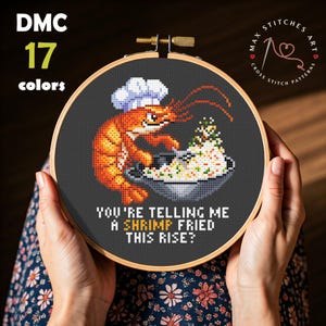 May include: Cross-stitch embroidery in a wooden hoop featuring a chef shrimp with a white hat cooking fried rice. The text reads "YOU'RE TELLING ME A SHRIMP FRIED THIS RISE?" with "SHRIMP FRIED" in bold. The design uses 17 colors.