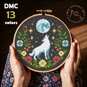 Peut inclure: Un motif de point de croix représentant un loup blanc hurlant à la lune avec une bordure florale. Le motif est encadré dans un cercle de broderie noir et tenu par les mains d'une personne. Le texte "MAX STITCHES ART CROSS STITCH PATTERNS" est visible dans le coin supérieur droit de l'image.