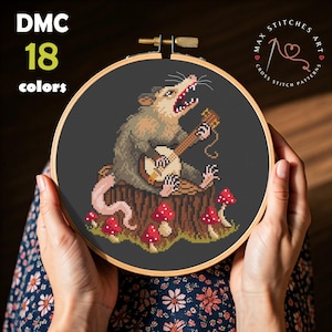 Op de afbeelding: Kruissteekpatroon met een opossum die banjo speelt op een boomstronk, omringd door rode paddenstoelen. Het ontwerp staat tegen een donkergrijze achtergrond in een houten borduurring. Tekst luidt "DMC 18 kleuren" en "MAX STITCHES ART CROSS STITCH PATTERNS".