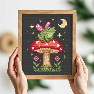 May include: Cross stitch pattern of a green frog wearing pink fairy wings sitting on a red and white mushroom with a yellow stem. The mushroom is surrounded by green leaves and small pink flowers. A crescent moon and stars are in the background.