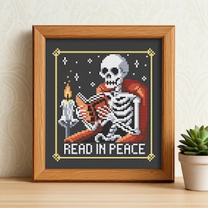 May include: A framed cross-stitch design featuring a skeleton reading a book by candlelight. The skeleton is sitting in a chair and the text reads "Read in Peace".