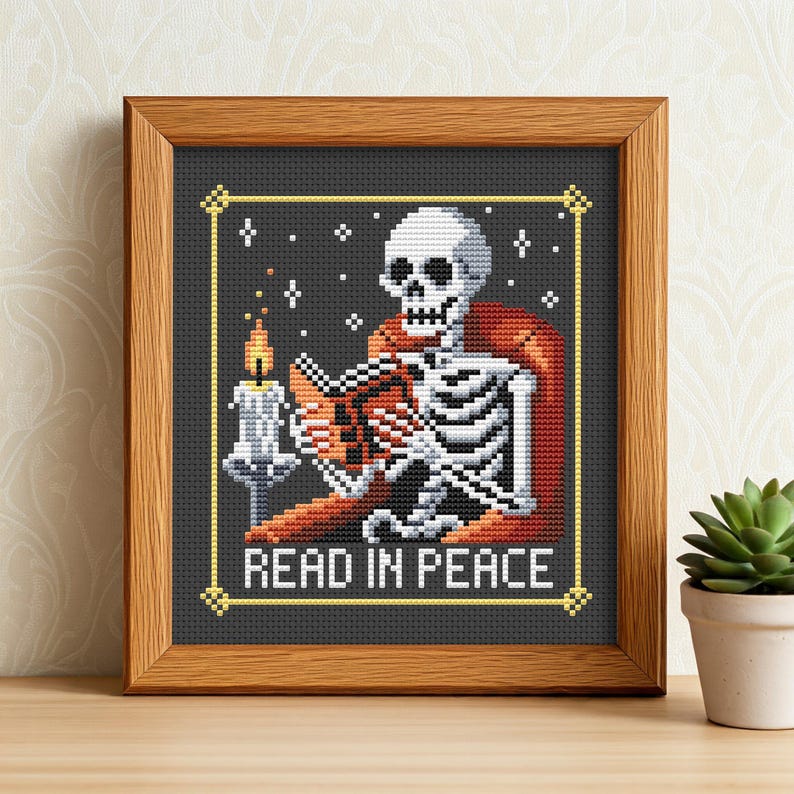 May include: A framed cross-stitch design featuring a skeleton reading a book by candlelight. The skeleton is sitting in a chair and the text reads "Read in Peace".