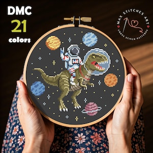 Puede incluir: Bordado de punto de cruz de un astronauta montando un dinosaurio por el espacio, rodeado de planetas. El diseño está cosido sobre tela negra y enmarcado en un aro de madera. El texto "DMC 21 colores" está en la esquina superior izquierda.