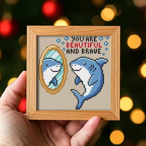 May include: A framed cross-stitch featuring a blue shark looking at its reflection in a mirror. The mirror and shark are surrounded by the words "YOU ARE BEAUTIFUL AND BRAVE" in red and blue. The frame is made of light brown wood.