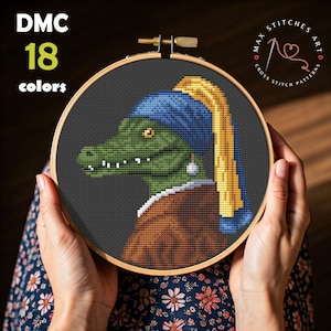 May include: Cross-stitch pattern featuring a green alligator in a blue and yellow head covering and a pearl earring, reminiscent of Vermeer's "Girl with a Pearl Earring." The text "DMC 18 colors" is visible. The cross-stitch is held in a wooden hoop.