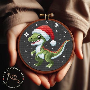 May include: A cross stitch pattern featuring a green dinosaur wearing a red Santa hat with white trim. The dinosaur is standing on a black background with white snowflakes.
