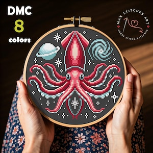May include: Cross stitch pattern of a red squid in space with a galaxy, stars, and a planet in the background. The pattern is in a round embroidery hoop. The text "Max Stitches Art Cross Stitch Patterns" is on the top right corner of the hoop.