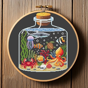 May include: Cross-stitch embroidery of an ocean scene inside a glass bottle. The design features a shark, jellyfish, octopus, crab, and various fish and coral. The embroidery is set in a wooden hoop.