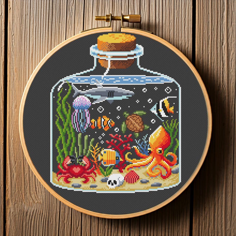 May include: Cross-stitch embroidery of an ocean scene inside a glass bottle. The design features a shark, jellyfish, octopus, crab, and various fish and coral. The embroidery is set in a wooden hoop.