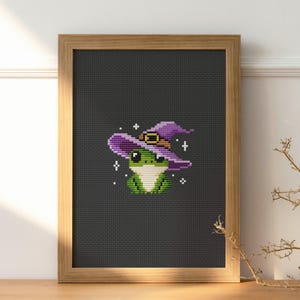 May include: Cross-stitch art of a green frog wearing a purple witch hat with a gold buckle. The frog is centered on a black background, framed in a light brown wooden frame. White star accents surround the frog.