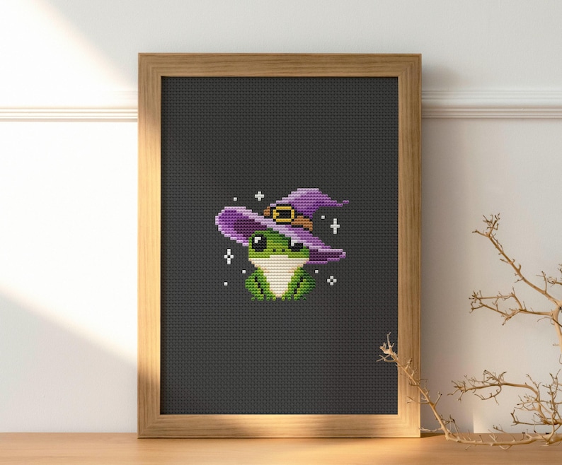 May include: Cross-stitch art of a green frog wearing a purple witch hat with a gold buckle. The frog is centered on a black background, framed in a light brown wooden frame. White star accents surround the frog.