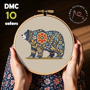 May include: A cross stitch pattern of a bear with floral details. The pattern is completed and framed in a wooden hoop. The pattern uses 10 colors of thread. The text "Max Stitches Art Cross Stitch Patterns" is visible in the upper right corner of the image.