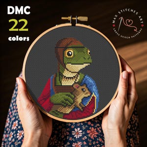 May include: Cross-stitch art in a wooden hoop featuring a green dinosaur-like creature holding a small brown animal. The design uses 22 colors and includes text that reads "DMC 22 colors" and "Max Stitches Art".