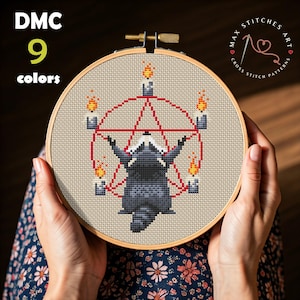 May include: Cross-stitch embroidery in a wooden hoop featuring a raccoon within a red pentagram, surrounded by candles with flames. The text "DMC 9 colors" is at the top left. The design is held by hands.
