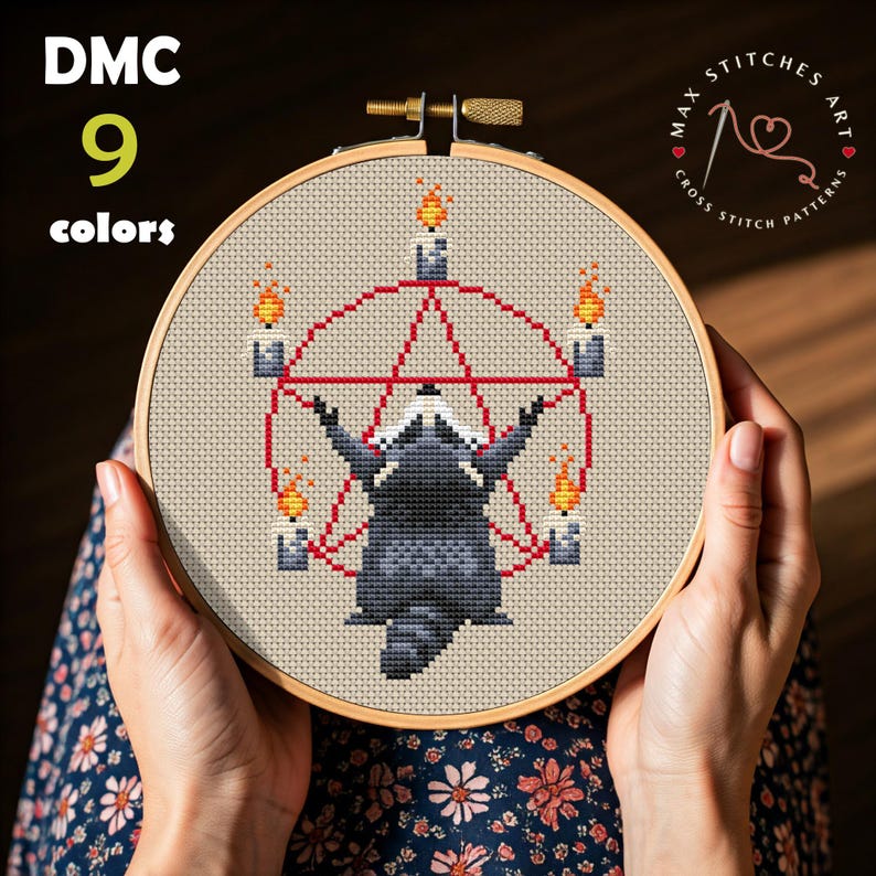 May include: Cross-stitch embroidery in a wooden hoop featuring a raccoon within a red pentagram, surrounded by candles with flames. The text "DMC 9 colors" is at the top left. The design is held by hands.