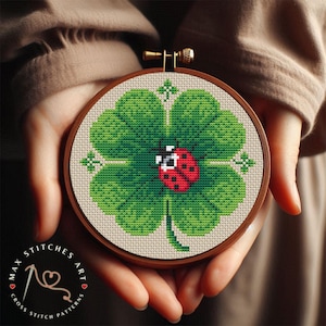 May include: A cross stitch pattern of a four-leaf clover with a red ladybug on it. The pattern is framed in a wooden hoop and held in someone's hands.