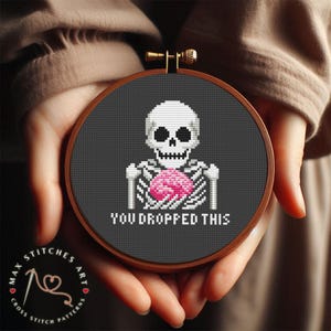 Puede incluir: Un patrón de punto de cruz en blanco y negro de un esqueleto sosteniendo un cerebro rosa con el texto "You dropped this".