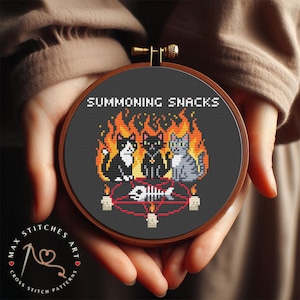May include: A circular wooden embroidery hoop containing a cross-stitch pattern on dark fabric. The design features three cats sitting before a red pentagram with a fish skeleton, surrounded by orange flames and the text SUMMONING SNACKS.