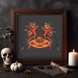 Puede incluir: Imagen de punto de cruz en un marco de madera oscura. La imagen presenta tres mapaches alrededor de una hamburguesa sobre una plataforma circular brillante. Los mapaches son de color naranja y negro, con un fondo gris oscuro. Una calavera, una calabaza y una vela en primer plano.