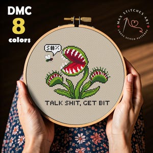 May include: A circular wooden embroidery hoop containing a cross-stitch design of a green Venus flytrap with a red interior and sharp white teeth. A small fly is nearby with a speech bubble containing the text S#%. The text TALK SHIT, GET BIT is below.
