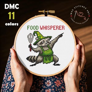 May include: Cross-stitch pattern featuring a raccoon chef with the text "FOOD WHISPERER." The raccoon wears a green chef's hat and apron, holding a whisk. The design is in a wooden embroidery hoop, with "DMC 11 colors" text.