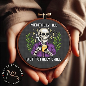 Puede incluir: Un bastidor de bordado de punto de cruz con un diseño de esqueleto con camisa morada y taza amarilla. El texto dice "MENTALLY ILL BUT TOTALLY CHILL". Fondo gris oscuro con detalles verdes.