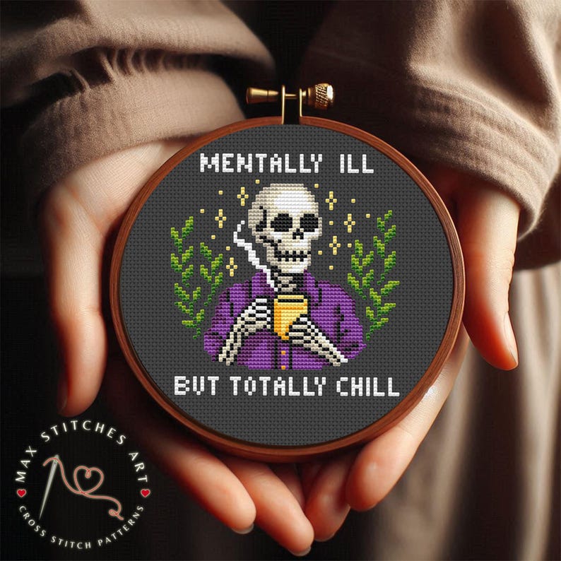 May include: A cross-stitch embroidery hoop with a skeleton design. The skeleton wears a purple shirt and holds a yellow cup, with the text "MENTALLY ILL BUT TOTALLY CHILL". The background is dark grey with green foliage accents.