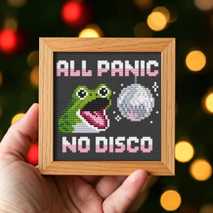 May include: A framed cross-stitch with a green frog, a disco ball, and the text "ALL PANIC NO DISCO" in pink and white. The design is on a dark grey background, framed in light brown wood. The artwork is held in a light brown wooden frame.