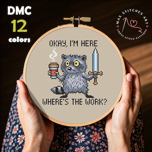 May include: A cross-stitch hoop featuring a raccoon holding a coffee cup and a sword, with the text "OKAY, I'M HERE. WHERE'S THE WORK?". The design includes the text "DMC 12 colors" and "MAX STITCHES ART".