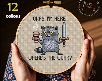 Pode incluir: Um bordado em ponto cruz num bastidor, apresentando um guaxinim com uma chávena de café e uma espada, com o texto "OKAY, I'M HERE. WHERE'S THE WORK?". O design inclui "DMC 12 cores" e "MAX STITCHES ART".