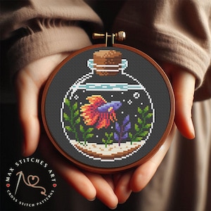 Op de afbeelding: Kruissteekpatroon van een betta-vis in een glazen pot met een kurken stop. De vis is oranje, rood en blauw met groene en paarse planten in de pot.