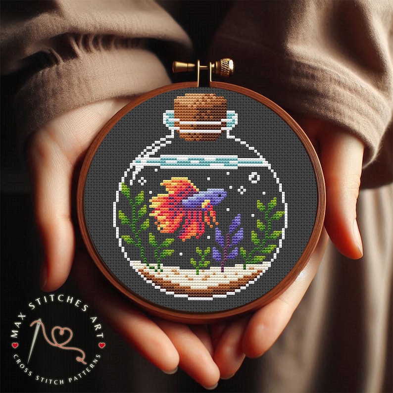 May include: Cross stitch pattern of a betta fish in a glass jar with a cork stopper. The fish is orange, red, and blue with green and purple plants in the jar.