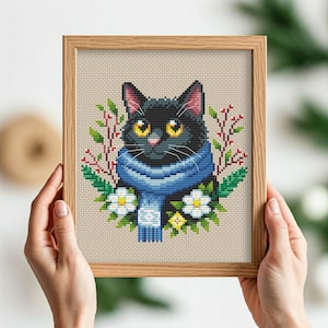 Op de afbeelding: Borduurpatroon van een zwarte kat met een blauwe sjaal met witte bloemen eromheen.