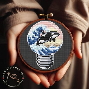 Könnte beinhalten: Ein Kreuzstichmuster einer Glühbirne mit einem schwarz-weißen Orca-Wal, der aus einer Welle springt, die sich im Inneren der Glühbirne befindet. Die Welle ist blau und weiß und der Hintergrund ist dunkelgrau.