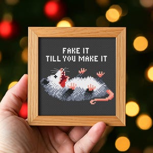 Könnte beinhalten: Ein gerahmtes Kreuzstichbild mit einem Opossum und dem Text "FAKE IT TILL YOU MAKE IT" in weißen Blockbuchstaben. Das Opossum ist grau, weiß und rosa, liegt auf dem Rücken und hat den Mund geöffnet. Der Rahmen ist aus hellem Holz.