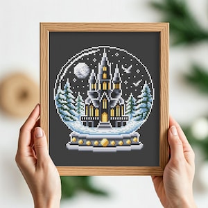 May include: A cross-stitch pattern of a snow globe with a black castle inside. The castle has yellow windows and is surrounded by trees. The snow globe is on a black background.