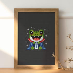 May include: Cross-stitch art of a green frog wearing a red and blue cape, framed in a light brown wooden frame. The frog has a wide, happy smile and is surrounded by white star-shaped sparkles. The background is a dark gray color.