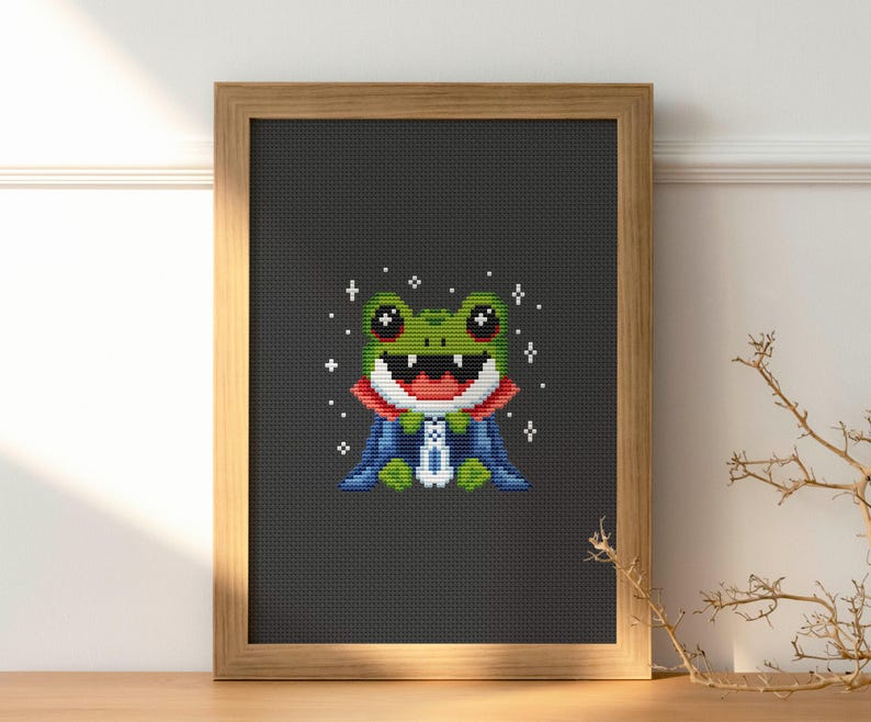 May include: Cross-stitch art of a green frog wearing a red and blue cape, framed in a light brown wooden frame. The frog has a wide, happy smile and is surrounded by white star-shaped sparkles. The background is a dark gray color.