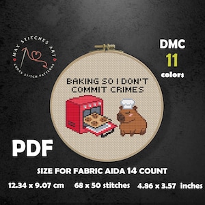 May include: Cross-stitch pattern in a wooden hoop featuring a red oven with cookies and a brown capybara. The text reads "BAKING SO I DON'T COMMIT CRIMES." Includes size information: 4.86 x 3.57 inches.
