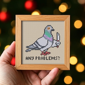 Könnte beinhalten: Kreuzstich-Kunstwerk eines Pigeons, der ein Messer hält, in einem hellbraunen Holzrahmen. Der Vogel ist grau, weiß und lila, mit dem Text "ANY PROBLEMS?" darunter. Der Hintergrund ist beige.