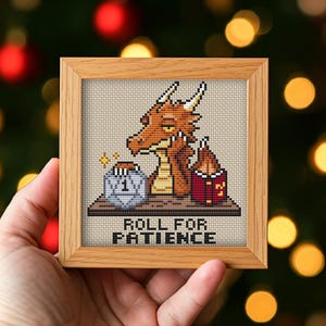 May include: A square wooden frame containing a cross-stitch embroidery of an orange dragon resting its head on its hand. The dragon sits behind a table with a twenty-sided die showing the number one and a red book. The text reads ROLL FOR PATIENCE.